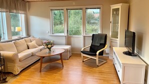 A living room with a sofa, armchair, coffee table, TV stand, and windows with a view of greenery. - Cosy Cottage in Dals-Ed, 3 Min Drive from Moose Ranch & Lakes (Ed)