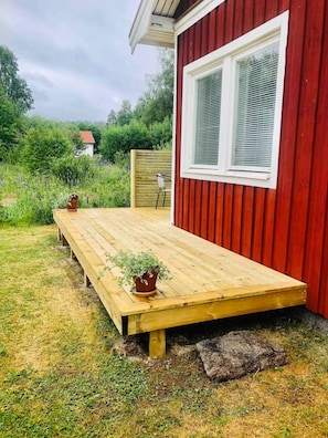 Eine Holzterrasse mit Blumentöpfen, ein rotes Gebäude mit einem Fenster und ein Holzzaun.