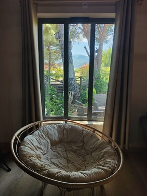 A room with a large window, a wicker chair, and a view of a garden. - Wooden house, country spirit, 10 minutes from the sea. (Saint-Génis-des-Fontaines)