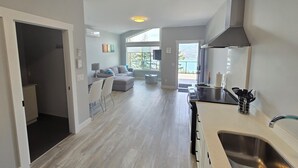 Cozinha moderna com pia de aço inoxidável, área de jantar com mesa e cadeiras, e sala de estar com sofá e vista para o exterior.
