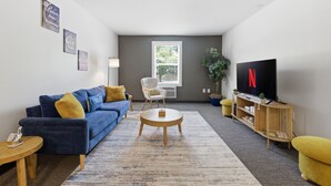 Un salon avec un canapé bleu, une table basse ronde, un téléviseur et une fenêtre avec des stores.