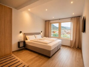 Une chambre moderne avec un grand lit, une table de chevet en bois, une commode, un téléviseur fixé au mur et une fenêtre avec des rideaux.