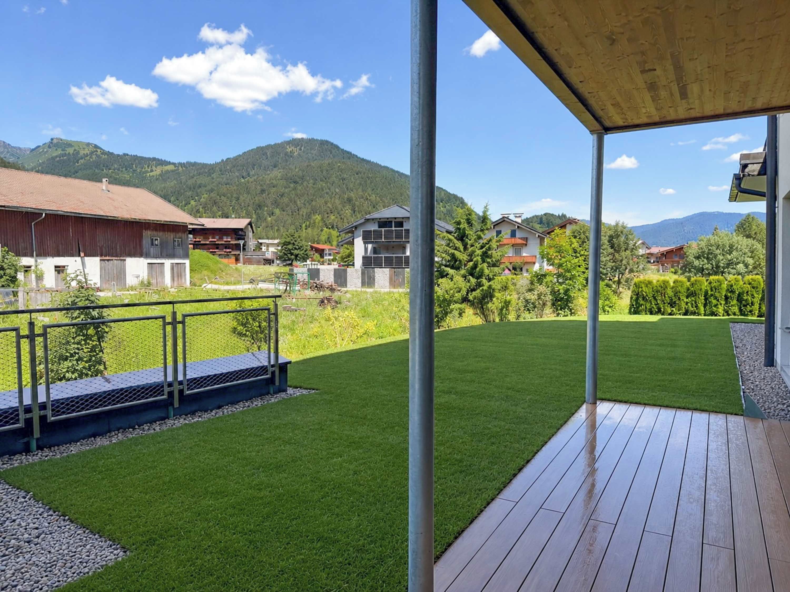 Ein Wohnhaus mit Holzterrasse, gepflegtem Rasen und Blick auf eine Berglandschaft.