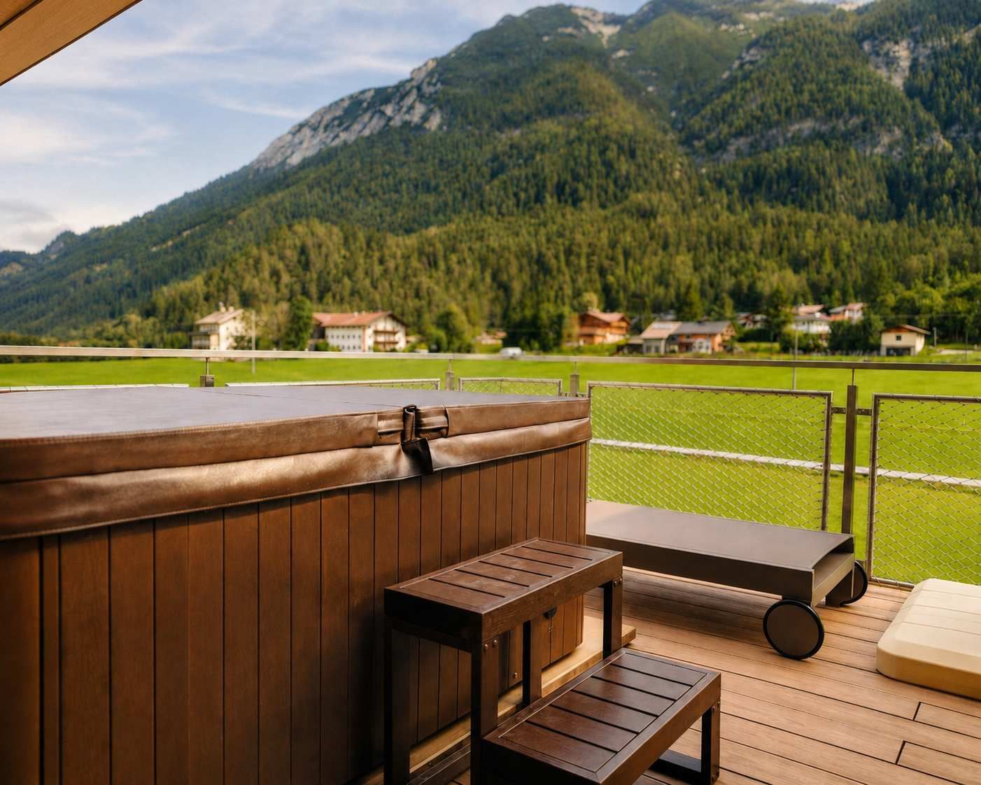 A wooden deck with a hot tub, a bench, and a lounge chair, overlooking a green field and mountains.