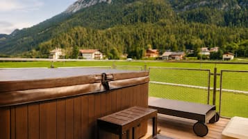 Une terrasse en bois avec un jacuzzi, un banc et un fauteuil de relaxation, offrant une vue sur un champ verdoyant et des montagnes.