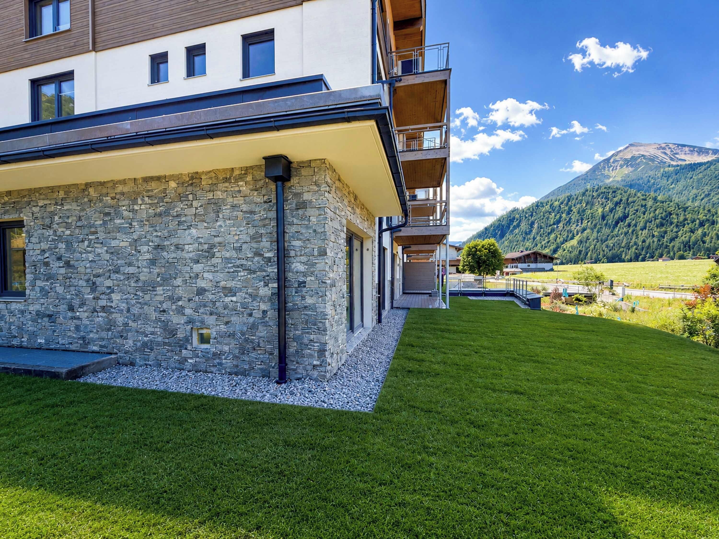 Ein modernes Gebäude mit Naturstein-Fassade, flachem Dach und einem Balkon mit Blick auf eine gepflegte Rasenfläche und eine gebirgige Landschaft.