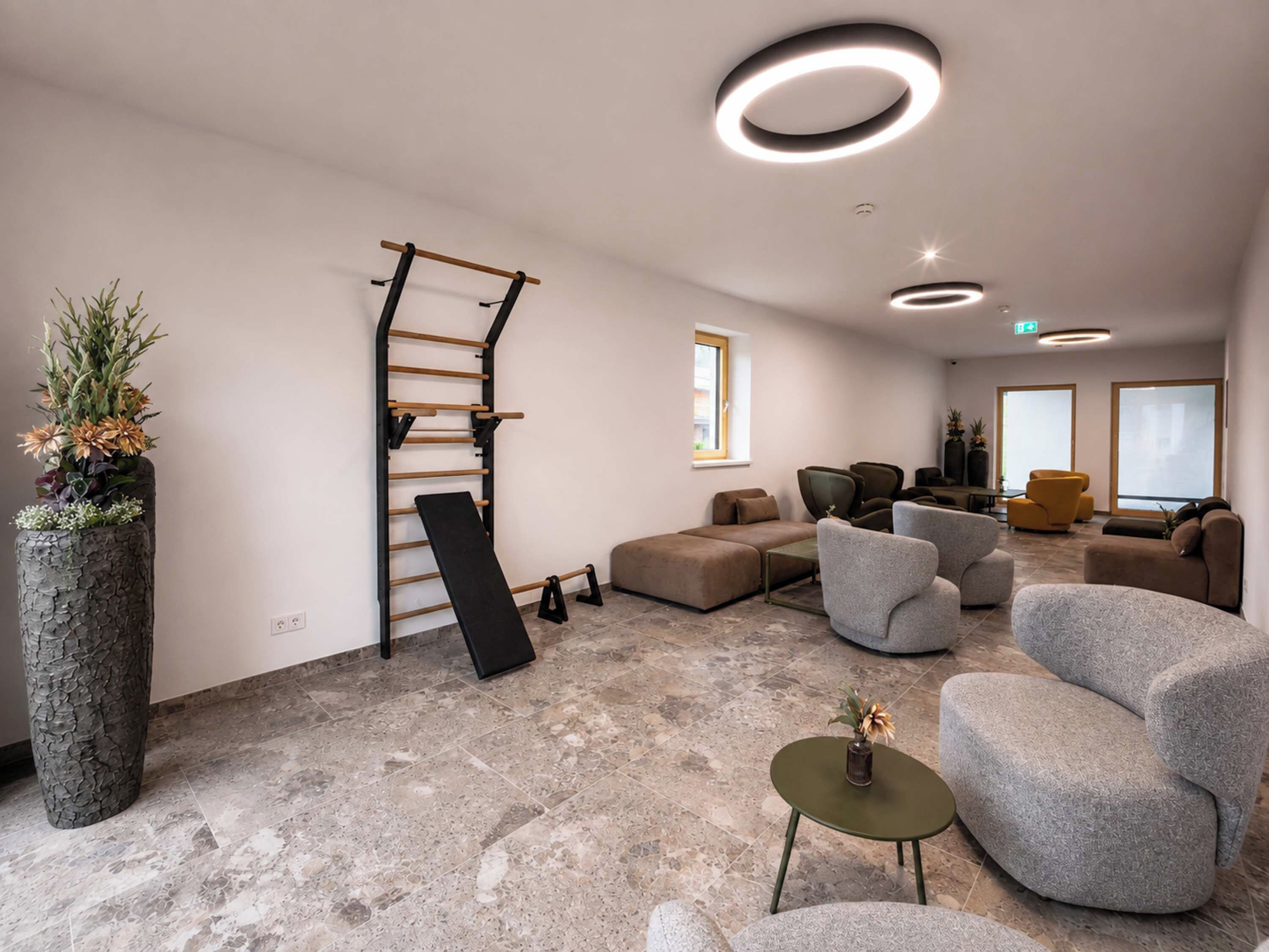 Un espace de détente moderne avec un sol en pierre, des canapés gris, un mur d’escalade en bois et une grande plante en pot.