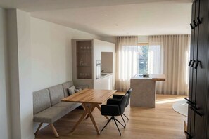 A modern living space with a wooden dining table, black chairs, a grey sofa, a kitchen area, and a large window with curtains.