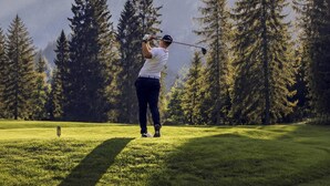 Uma pessoa jogando golfe em um campo com árvores altas e montanhas ao fundo.