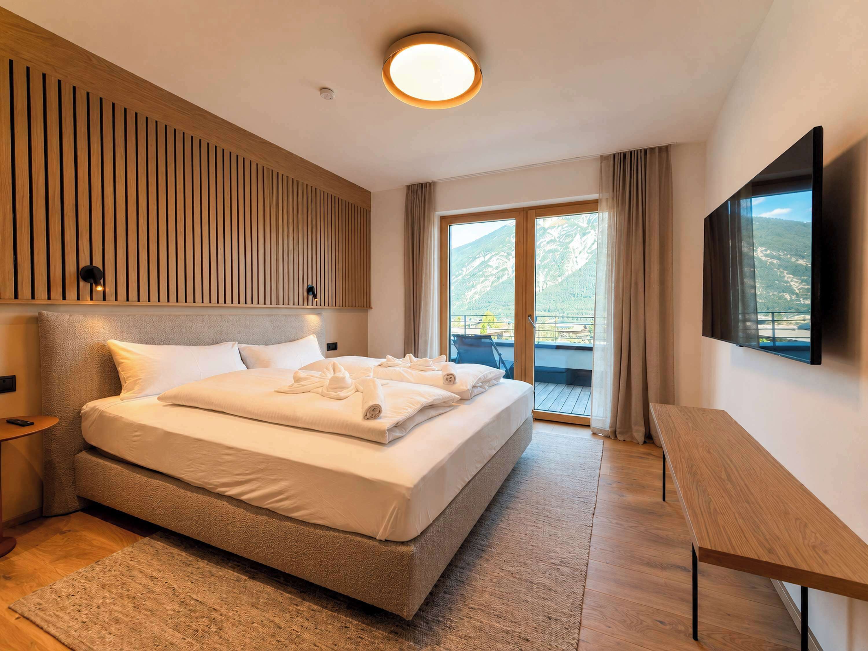 Ein Hotelzimmer mit einem großen Bett, einem Flachbildfernseher, einem Holz-Kopfteil, einem Balkon mit Bergblick und einem kleinen Tisch.