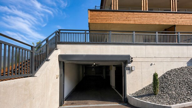 Entrada de um edifício moderno com rampa de concreto, guarda-corpos metálicos e uma área de cascalho com uma pequena árvore.