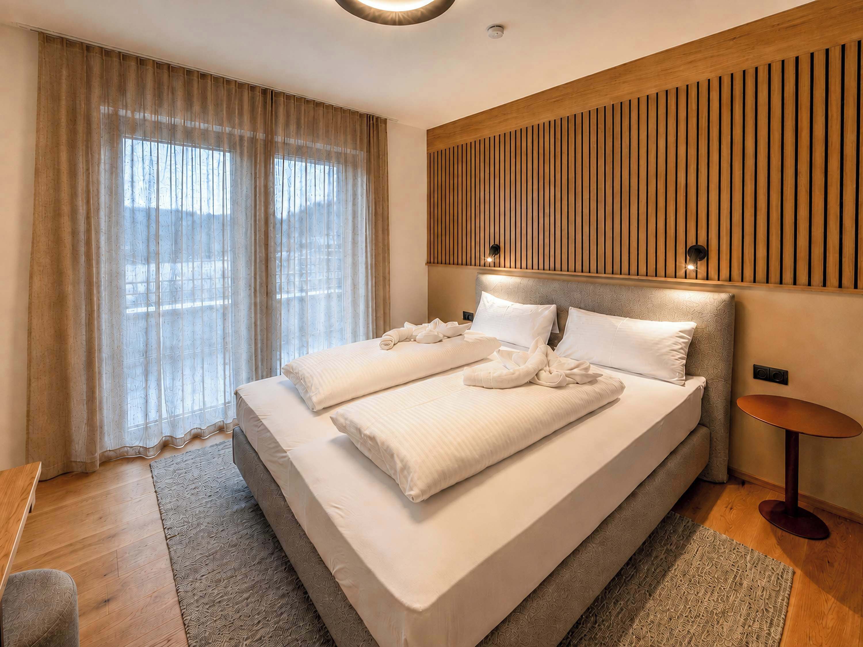 A modern hotel room with a large bed, a wooden headboard, a small round table, and a view through the window.