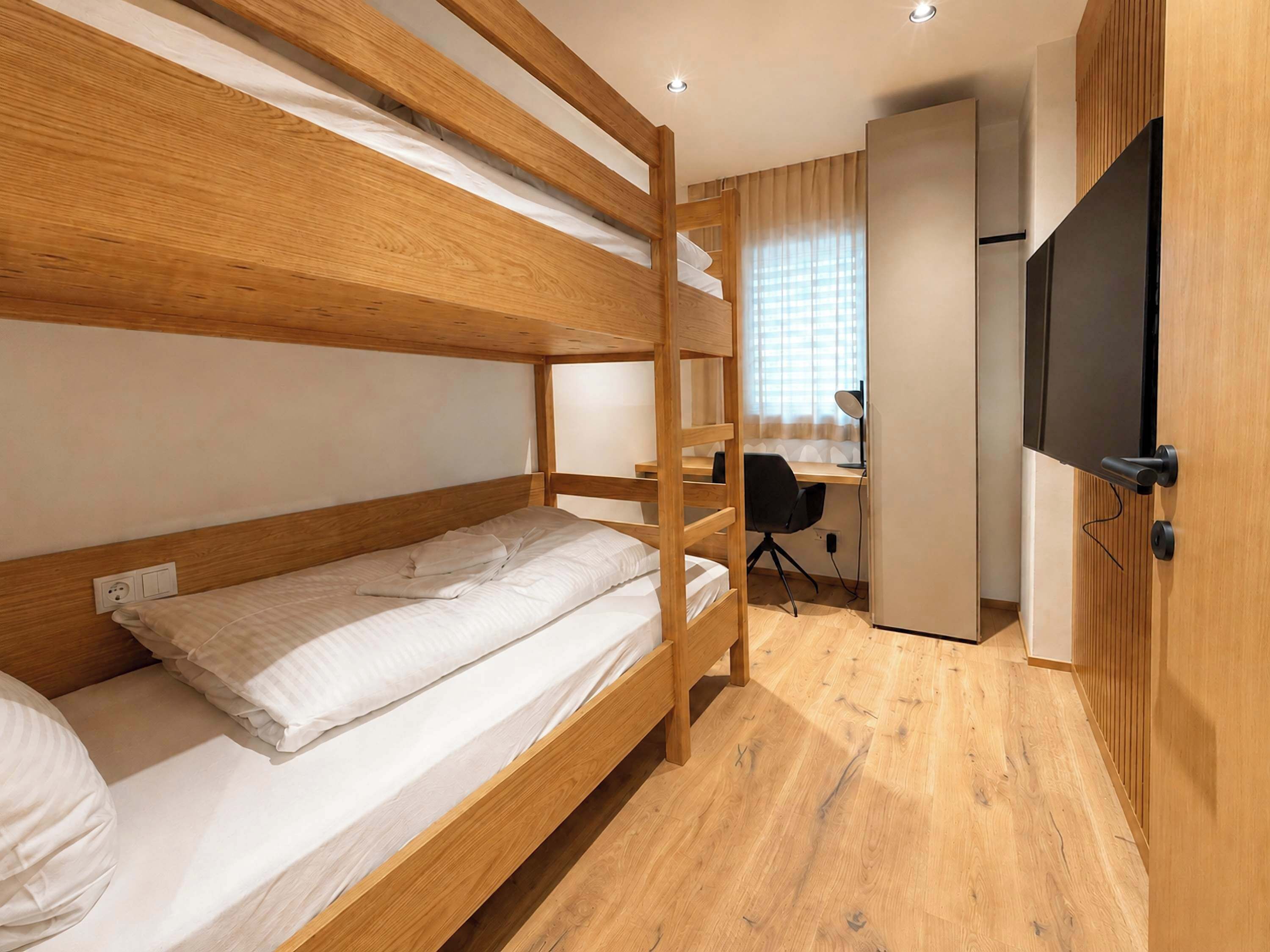 Ein Stockbett mit Schreibtisch und Stuhl, einem Fernseher und einer Steckdose in einem Zimmer mit Holzboden und einem Fenster mit Jalousien.