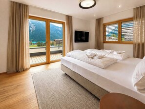 Habitación de hotel con cama, televisión de pantalla plana, balcón con vistas a la montaña y suelo de madera.