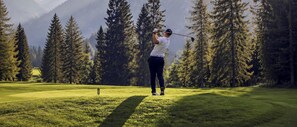 A person playing golf on a course with a backdrop of tall trees and mountains.