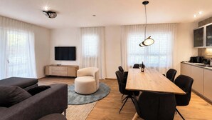 Een moderne woonkamer met een houten eettafel, zwarte stoelen, een grijze bank en een flatscreen televisie aan de muur.
