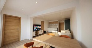 Ein modernes Wohnzimmer mit einem Holzesstisch, orangefarbenen Sesseln, einem Fernseher und einer Küche im Hintergrund.