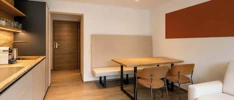 A modern kitchen with a wooden countertop, a built-in oven, and a sink. There is a dining area with a wooden table and four chairs. A sofa is placed against the wall, and a door leads to another room.