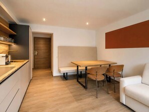 Eine moderne Küche mit einer Holzausziehtafel, einem eingebauten Ofen und einem Spülbecken. Ein Essbereich mit einem Holztisch und vier Stühlen lädt zum Verweilen ein. Ein Sofa steht an der Wand, und eine Tür führt in einen weiteren Raum.