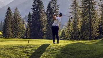 Een persoon speelt golf op een veld, met hoge bomen en bergen op de achtergrond.