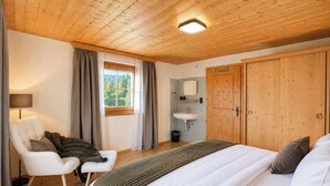 Ein Schlafzimmer mit Bett, Stuhl, Nachttisch, einem Fenster mit Vorhängen, einer Holzbalkendecke und einem Holzschrank.