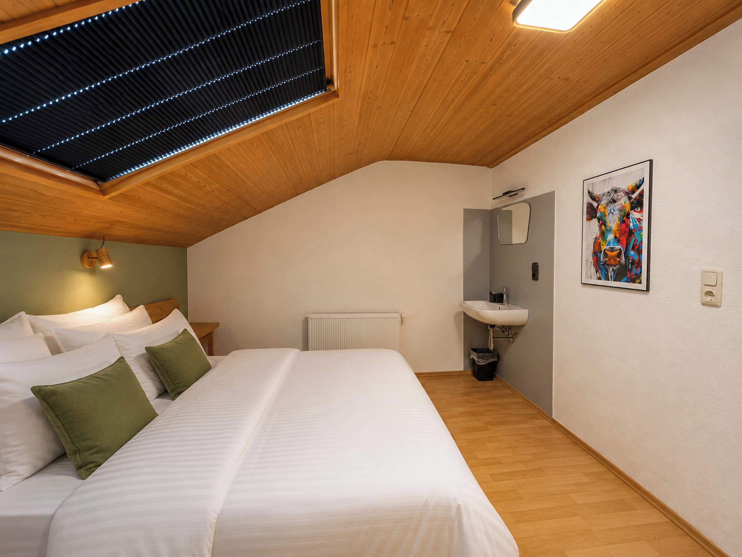 Une chambre avec un plafond en pente, un lit avec du linge de lit blanc, une table de chevet, un radiateur, un lavabo fixé au mur et une œuvre d’art encadrée.