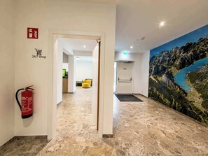 Eine Hotelhalle mit Feuerlöscher, Schild für einen Kiosk, der rund um die Uhr geöffnet ist, und Wandgemälde einer Berglandschaft.
