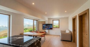A modern kitchen with a marble countertop, a dining area with chairs, a television mounted on the wall, and large windows offering a view of the mountains. - BASE Achensee Soul No1 – Private sauna (Achenkirch)