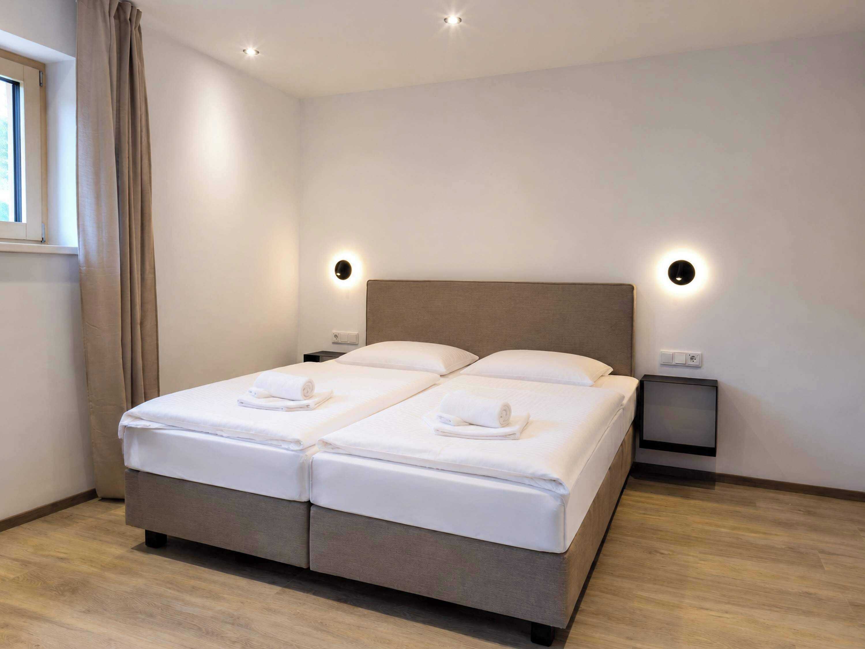 Un lit double avec deux oreillers et deux serviettes enroulées, des tables de chevet, une fenêtre avec des rideaux et des spots encastrés au plafond.