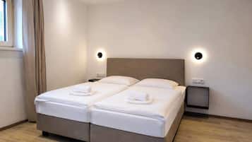 Un lit double avec deux oreillers et deux serviettes enroulées, des tables de chevet, une fenêtre avec des rideaux et des spots encastrés au plafond.