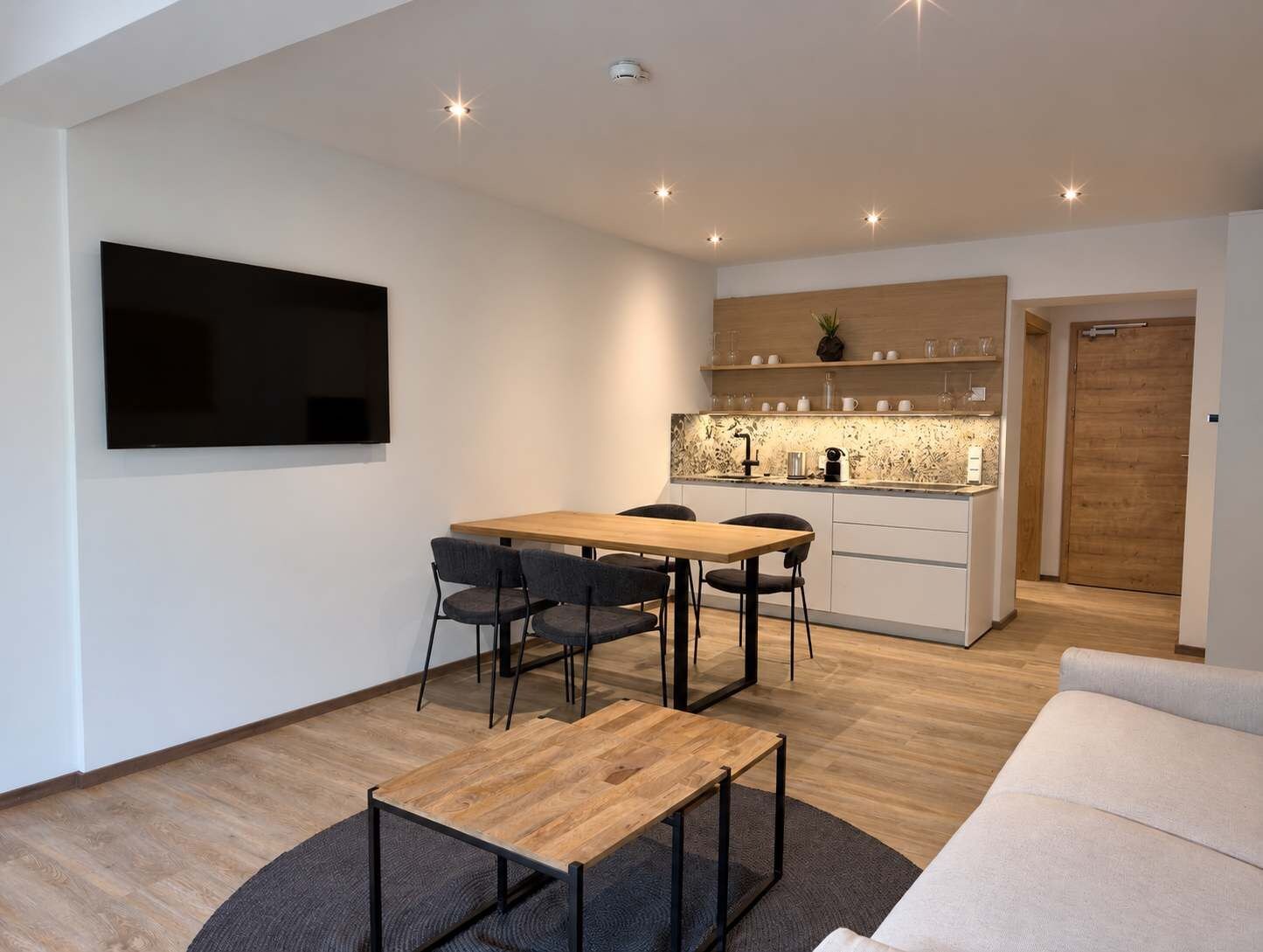 A modern living room with a wooden dining table, black chairs, a TV mounted on the wall, and a kitchen area with wooden cabinets and a countertop.