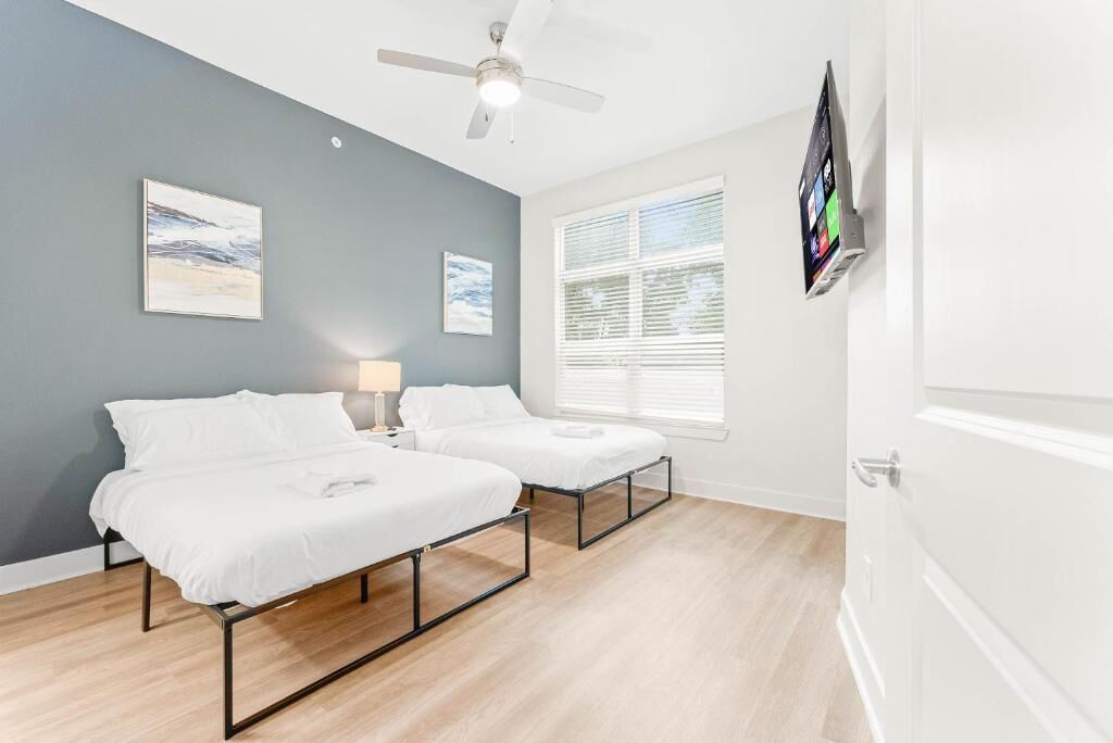 A room with two beds, a ceiling fan, a TV mounted on the wall, and a window with blinds.