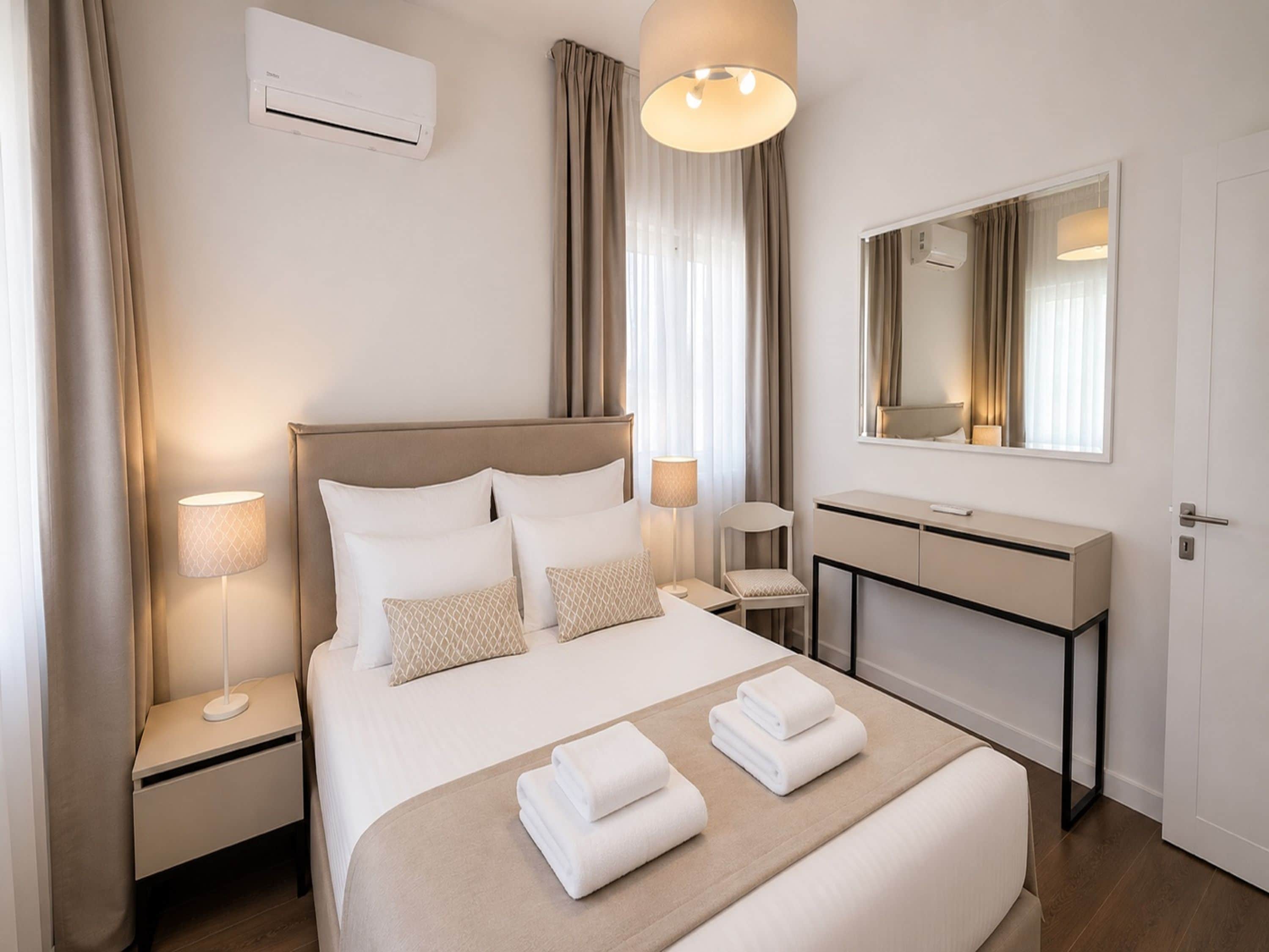 Una habitación de hotel con cama, lámparas de noche, escritorio, espejo y aire acondicionado.