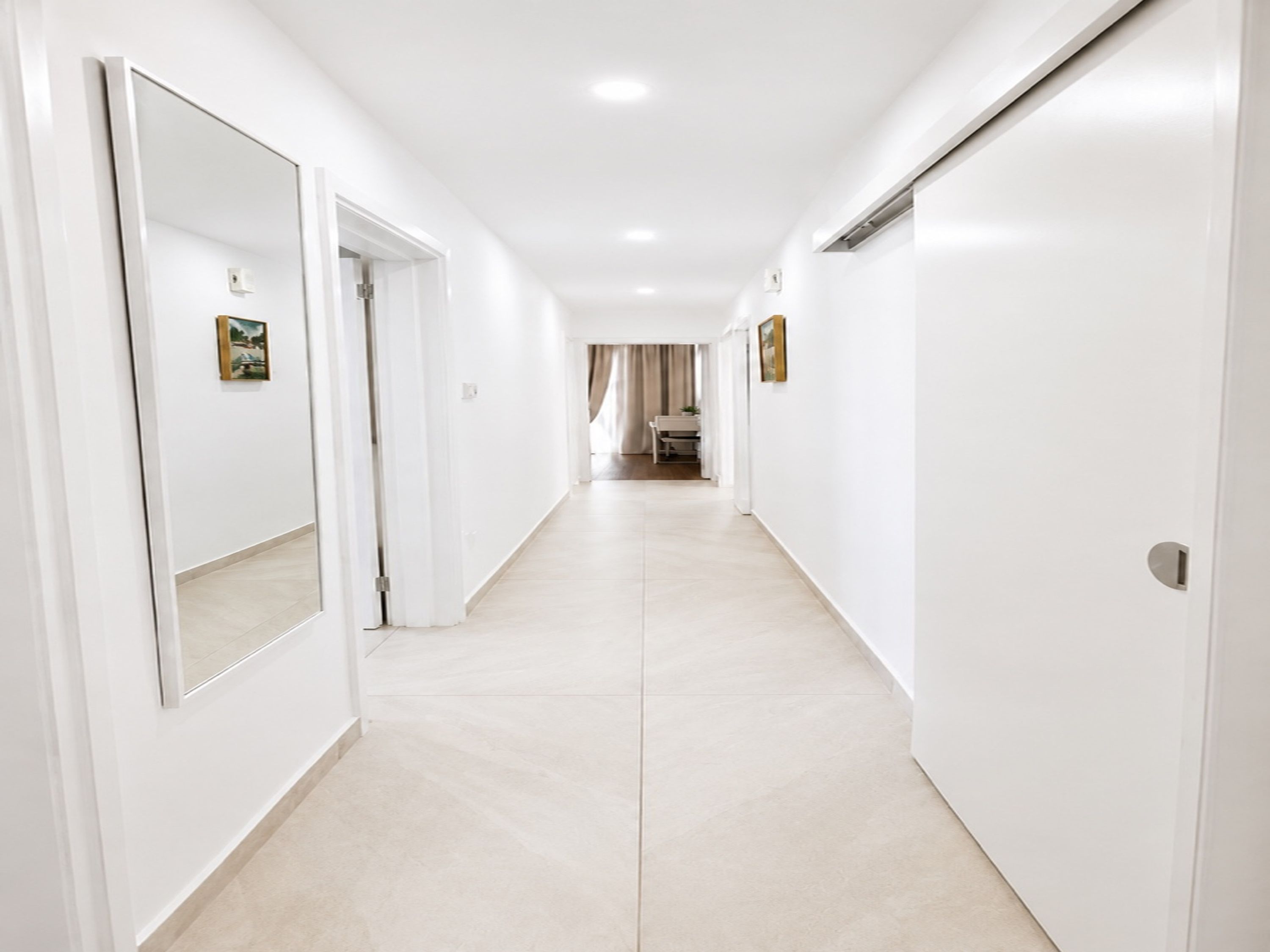 A long, narrow hallway with white walls and a tiled floor, leading to a room with a bed and a window.