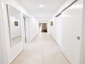Ein langer, schmaler Flur mit weißen Wänden und Fliesenboden, der zu einem Zimmer mit Bett und Fenster führt.