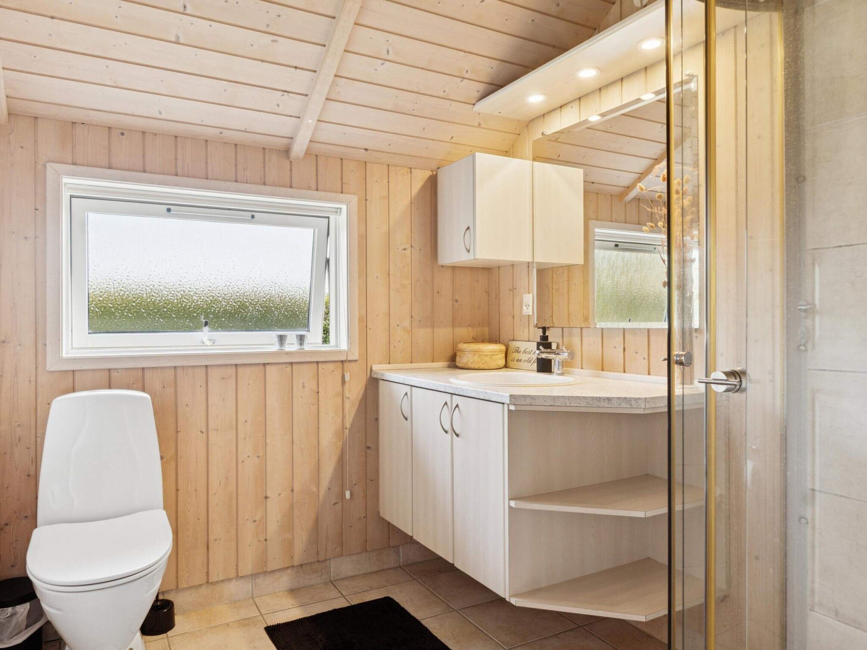 A bathroom with wooden walls, a white toilet, a window, a mirror, and a sink with a cabinet.