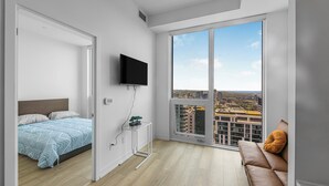 A modern bedroom with a bed, a TV, a small table with a plant, and a sofa with a cushion. - Saint Suites - Penthouse 2Bed+2Bath High Rise Condo (Toronto)