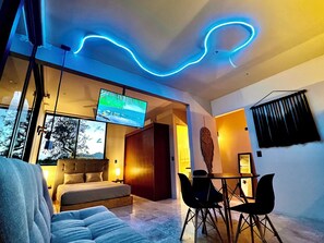 Ein modernes Wohnzimmer mit einer Couch, einem an der Wand befestigten Fernseher, einem Essbereich mit Stühlen und einem dekorativen Wandbild.