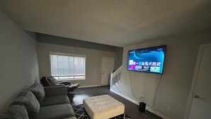 A living room with a sectional sofa, ottoman, and a flat-screen TV mounted on the wall. -  Modern Vinings Loft 1BR Retreat Near The Battery & Atlanta (Atlanta)