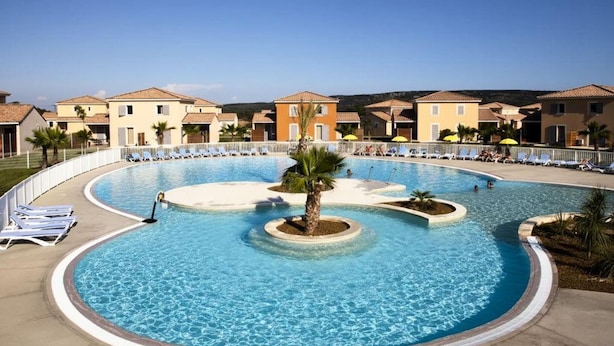Una amplia piscina con sillas de descanso y palmeras, rodeada de edificios con techos de tejas.
