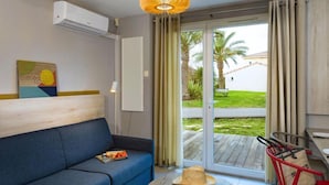 Una habitación con un sofá azul, una mesa redonda con un sombrero y un libro, una silla roja y vistas a un jardín por una puerta corredera.