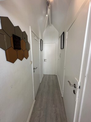 Ein Flur mit Holzboden, weißen Wänden und sechseckigen Wanddekorationen. Auf jeder Seite befinden sich je zwei Türen, eine mit Türgriff und eine mit Hebel.