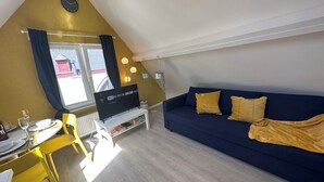 Una sala de estar acogedora con un sofá azul, una mesa de centro blanca y un televisor sobre un mueble bajo. Hay un comedor con sillas amarillas y una mesa dispuesta para dos personas.