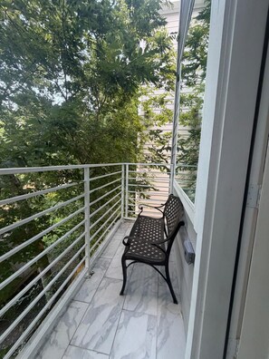 Ein Balkon mit Metallgeländer, einer Bank und Blick auf Bäume und ein Gebäude.