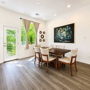 A dining area with a wooden table and chairs, a painting on the wall, and a door leading to a balcony with a view of greenery. - NRG Summer Haven Stay | 3 Stories | Sleeps 6 (Houston)