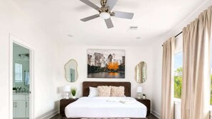 Quarto com uma cama grande, um ventilador de teto, uma pintura com paisagem urbana, um banco e vista para o exterior.