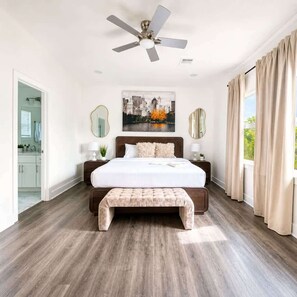 A bedroom with a large bed, a ceiling fan, a cityscape painting, a bench, and a view of the outdoors. - NRG Summer Haven Stay | 3 Stories | Sleeps 6 (Houston)