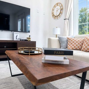 A modern living room with a wooden coffee table, a television, a bookshelf with books, a gold-framed clock, and a view through a window. - NRG Summer Haven Stay | 3 Stories | Sleeps 6 (Houston)