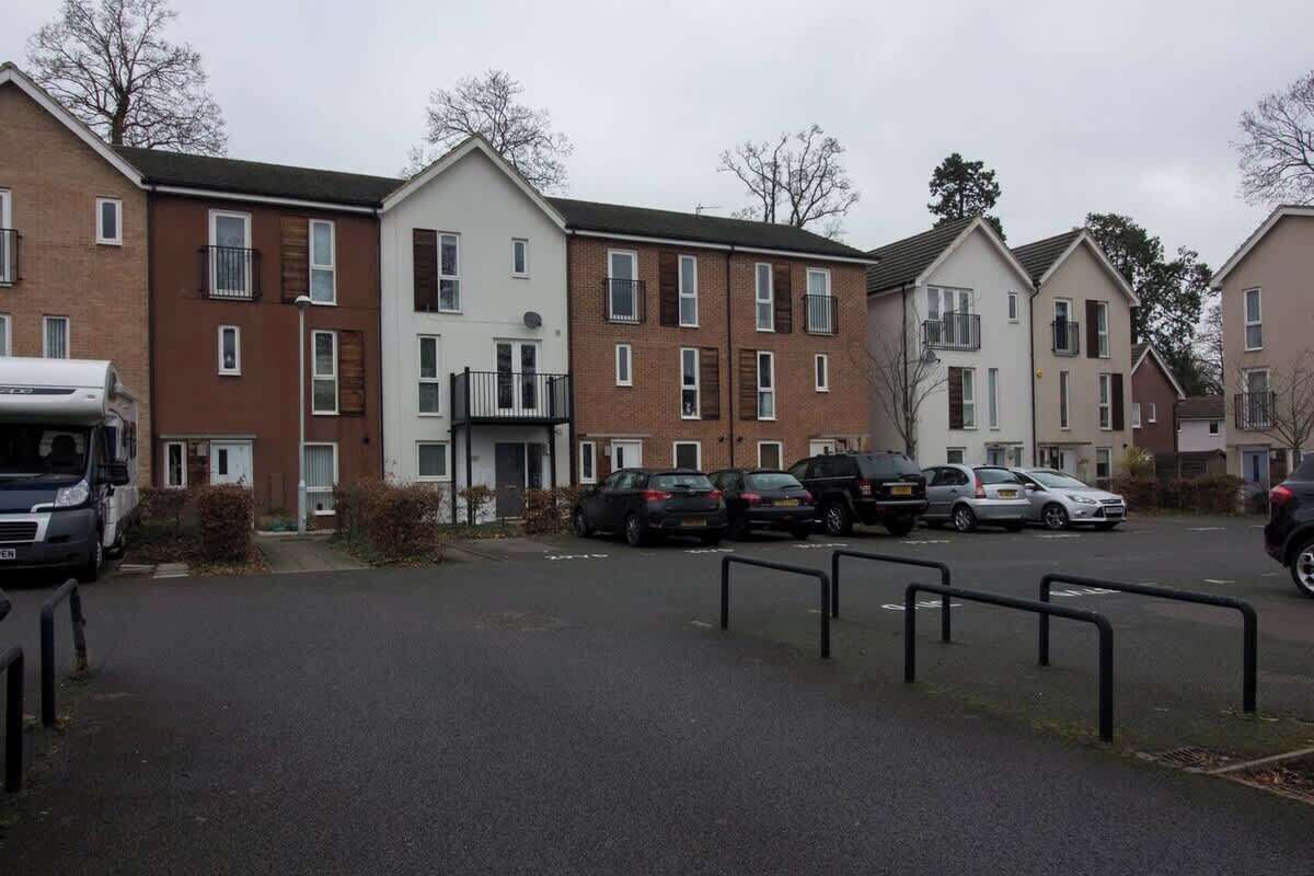 Una zona residencial con varias casas de dos pisos, un estacionamiento con varios automóviles y una camioneta blanca estacionada al costado.