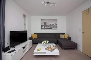 A modern living room with a flat-screen TV, a sofa, a coffee table with fruits and a plate, and a cityscape painting on the wall. - Beautiful 4 Bedroom House Berkshire - Free Parking (Bracknell)